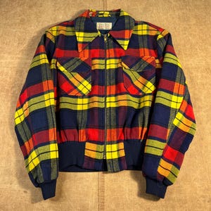 Könnte beinhalten: Eine Vintage-Jacke mit Reißverschluss und einem lebendigen rot, gelb und marineblauen Karomuster. Die Jacke hat zwei Vordertaschen, einen gerippten Saum und Bündchen sowie einen Kragen. Es gibt ein Etikett am Hals.