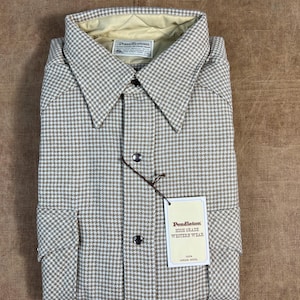 Op de afbeelding: Een gevouwen overhemd met een beige en wit pied-de-poule patroon, met een kraag en twee voorzakken. Het overhemd heeft een label met de tekst "Pendleton HIGH GRADE WESTERN WEAR 100% VIRGIN WOOL."
