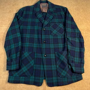 May include: A vintage plaid jacket in shades of navy blue and teal green. The jacket features a classic collar, three black buttons, and two large front pockets. The jacket has a tailored fit and is made of a thick, woven fabric.