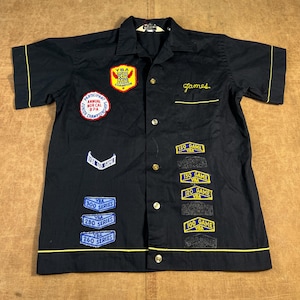 May include: Black bowling shirt with yellow trim and patches. Patches include "YBA", "Participant", and "Series" labels. The name "James" is embroidered on the chest. The shirt has short sleeves and a button-down front.