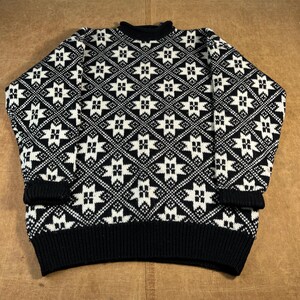 Könnte beinhalten: Ein schwarz-weiß gemusterter Pullover mit Rundhalsausschnitt und langen Ärmeln. Der Pullover hat ein sich wiederholendes geometrisches Design aus weißen sternartigen Formen in einem Rautenmuster. Die Bündchen, der Kragen und der Saum sind schwarz.