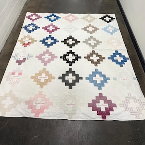 Könnte beinhalten: Eine weiße Steppdecke mit einem sich wiederholenden Muster aus kreuzförmigen Blöcken in verschiedenen Farben wie Rosa, Blau, Braun, Schwarz und Beige. Jeder farbige Block enthält mittig gestickten Text, der in einem Raster angeordnet ist.