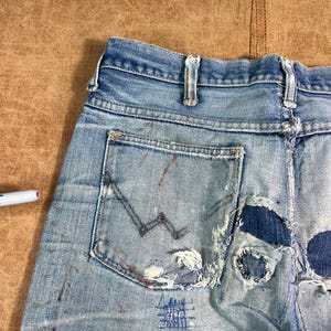 May include: Light blue denim jeans with distressed details, including tears and patches. The jeans feature a back pocket with a stitched design and paint splatters. The overall look is vintage and worn.