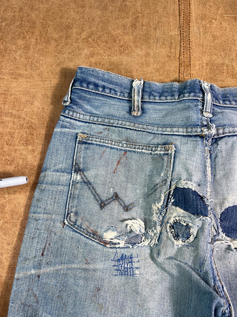 May include: Light blue denim jeans with distressed details, including tears and patches. The jeans feature a back pocket with a stitched design and paint splatters. The overall look is vintage and worn.