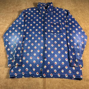 May include: A blue button-down shirt featuring a repeating pattern of colourful, abstract shapes. The long-sleeved shirt has a single chest pocket and a classic collar. The pattern includes red, yellow, and white accents on a blue background.