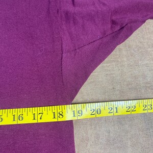May include: Close-up of a deep purple fabric with a yellow measuring tape. The tape shows measurements from 16 to 23 inches. The fabric appears to be a t-shirt or similar garment.