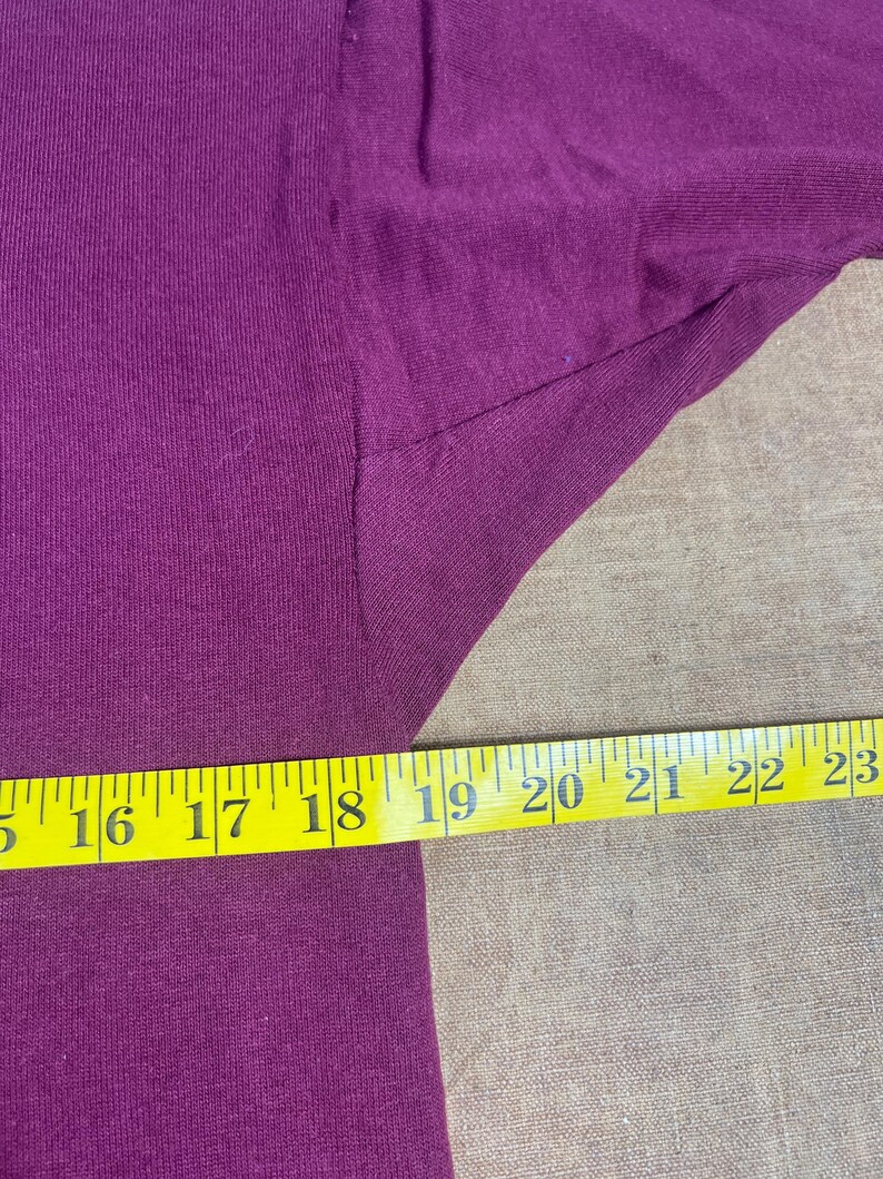 May include: Close-up of a deep purple fabric with a yellow measuring tape. The tape shows measurements from 16 to 23 inches. The fabric appears to be a t-shirt or similar garment.