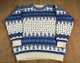 May include: Cream-colored sweater with a blue and white geometric pattern. The sweater features a crew neck, long sleeves, and ribbed cuffs and hem. The design includes various shapes and patterns in shades of blue, with accents of red and green.