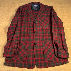 Könnte beinhalten: Ein rot-grüner Blazer mit Karomuster, drei schwarzen Knöpfen und zwei großen Vordertaschen. Der Blazer hat einen Reverskragen und eine einzelne Brusttasche. Die Ärmel haben geknöpfte Manschetten. Das Kleidungsstück ist aus einem gewebten Stoff.