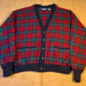 May include: A red, green, and navy blue plaid cardigan sweater with a V-neck, button closure, and two front pockets. The sweater has navy blue ribbed cuffs and hem.