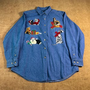 May include: A long-sleeved, button-down denim shirt with embroidered holiday-themed puppies and kittens. The shirt features various dog and cat breeds wearing Santa hats and festive decorations. The shirt is a medium blue wash.