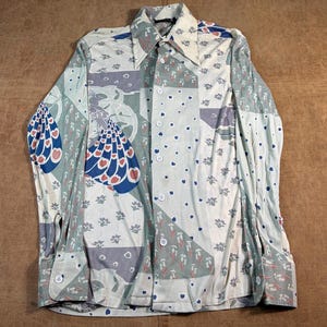 May include: A long-sleeve button-down shirt featuring a multi-panel patchwork design in shades of cream, sage green, and blue. The pattern includes stylized peacock motifs, small floral sprigs, and heart shapes, finished with white buttons.