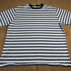 May include: A short-sleeved, striped t-shirt. The shirt features horizontal stripes in white, navy blue, and a lighter blue-grey. The neckline and sleeve cuffs are navy blue. The shirt is laid flat on a brown surface.