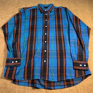 May include: A blue, black, and orange plaid shirt with a button-down front. The long-sleeved shirt features a classic collar and buttoned cuffs. The shirt is made of a woven fabric.
