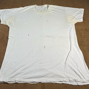 May include: A well-worn white t-shirt with a crew neck and short sleeves. The shirt displays signs of wear, including discolouration and small holes. The fabric appears to be cotton. The shirt is laid flat on a brown surface.