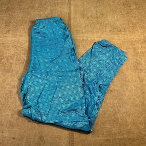 May include: Turquoise trousers with an elasticated waistband. The trousers feature a geometric pattern and are made of a shiny fabric. The trousers are folded on a brown surface.