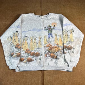 Puede incluir: Sudadera gris claro con botones con una escena de cosecha de otoño. El gráfico incluye un espantapájaros, calabazas, mazorcas de maíz y un carro de madera. La sudadera tiene cuello redondo y mangas largas.
