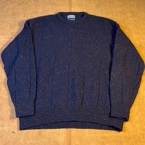 May include: A navy blue sweater with a textured diamond pattern. The sweater has a crew neck and ribbed cuffs and hem. The fabric has flecks of color throughout. A small tag is visible on the back of the neck.