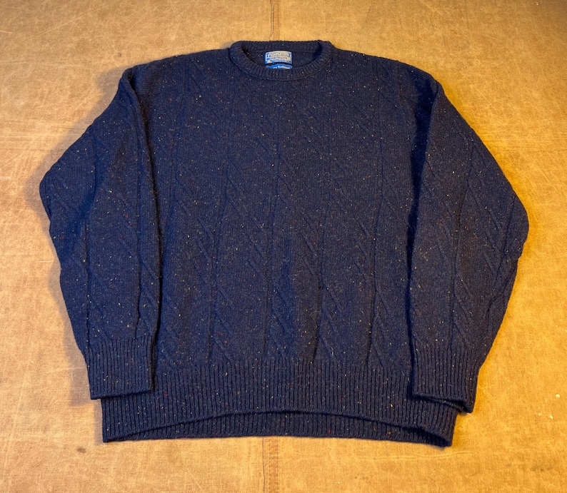 May include: A navy blue sweater with a textured diamond pattern. The sweater has a crew neck and ribbed cuffs and hem. The fabric has flecks of color throughout. A small tag is visible on the back of the neck.