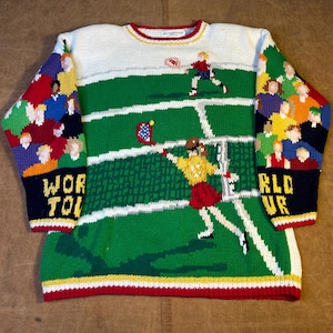 Könnte beinhalten: Ein Strickpullover mit einem Tennisplatz-Design. Der Pullover hat ein grünes Feld mit Tennisspielern, ein weißes Netz und Zuschauer. Die Ärmel tragen die Worte "WORLD TOUR" in Gelb und Weiß auf marineblauem Hintergrund.