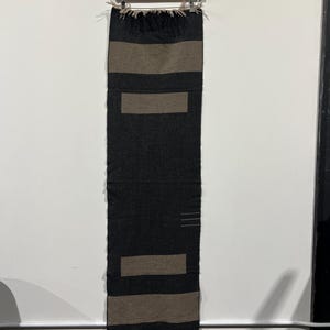 May include: A dark gray scarf with two horizontal tan rectangular designs. The scarf has a fringed edge and is hanging from a wooden hanger. The scarf is made of a soft material.