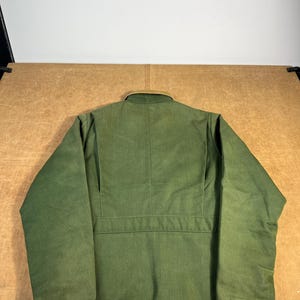 May include: A green jacket with a tan collar, viewed from the back. The jacket has a button closure and a horizontal seam across the back. The fabric appears to be a durable, possibly workwear-style material.