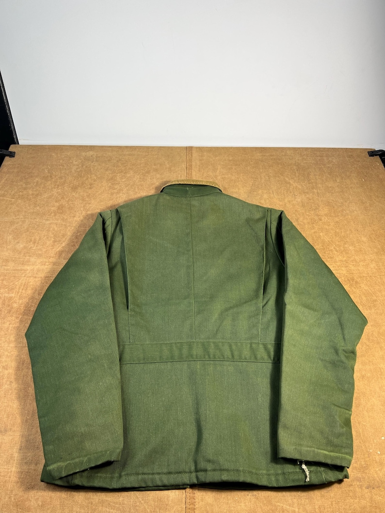 May include: A green jacket with a tan collar, viewed from the back. The jacket has a button closure and a horizontal seam across the back. The fabric appears to be a durable, possibly workwear-style material.