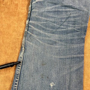 May include: Close-up of light blue denim jeans with visible wear and tear. The jeans show fading, creases, and a small hole near the bottom. A black pen rests on the edge of the jeans, providing a size reference.