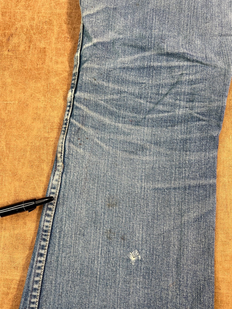 May include: Close-up of light blue denim jeans with visible wear and tear. The jeans show fading, creases, and a small hole near the bottom. A black pen rests on the edge of the jeans, providing a size reference.