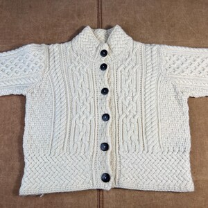 May include: Cream-colored, button-down cardigan sweater with a textured knit design. The sweater features a collared neckline and dark buttons. The knit pattern includes cable and diamond stitch details, creating a classic, cozy look. A timeless piece for layering.