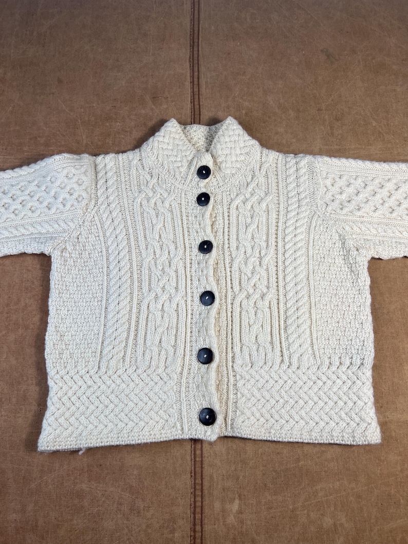 May include: Cream-colored, button-down cardigan sweater with a textured knit design. The sweater features a collared neckline and dark buttons. The knit pattern includes cable and diamond stitch details, creating a classic, cozy look. A timeless piece for layering.
