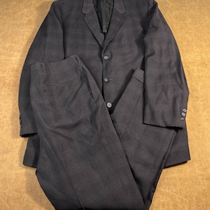 Pode incluir: Um fato cinza escuro, composto por um blazer de três botões e calças a condizer. O fato, feito de tecido texturizado, está sobre uma superfície bege. O blazer tem lapelas entalhadas e punhos abotoados. As calças têm um corte clássico e sob medida.