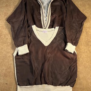 May include: Two-piece vintage brown athletic wear set. The top is a zip-up jacket with a cream-colored collar and trim. The bottom is a V-neck shirt with cream-colored trim at the neckline, cuffs, and hem. Both pieces have a ribbed texture.