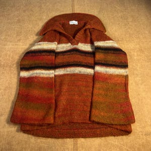 May include: A vintage, rust-colored knit sweater with a wide collar and striped design. The sweater features horizontal stripes in black, white, and various shades of orange and brown. The sleeves are flared, and the overall style is reminiscent of the 1970s.
