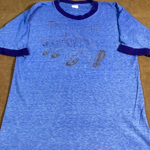 May include: Blue t-shirt with purple trim and the words "Keep on Pushin'!" above a graphic of cartoon figures walking. The shirt is a light blue color and has short sleeves.