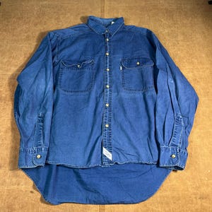 May include: A long-sleeved, blue denim shirt with two chest pockets and a button-down front. The shirt has a curved hem and buttoned cuffs. The fabric appears worn, with a small tag near the bottom.