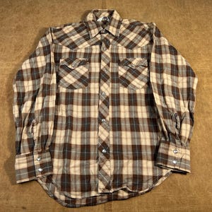 May include: A long-sleeved plaid shirt in shades of brown, beige, and blue. The shirt has two chest pockets, a button-down front, and buttoned cuffs. The fabric has a classic Western style.