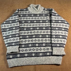 May include: A long-sleeve sweater featuring a high turtleneck collar and a repeating horizontal pattern of dark charcoal gray diamonds and small squares set against a textured, light heather gray knit background with ribbed cuffs and hem.