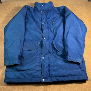 May include: A blue winter jacket with a high collar, multiple pockets, and a zipper closure. The jacket has a button-up front and a dark blue color. The sleeves have a dark blue cuff. The jacket is made of a durable material.