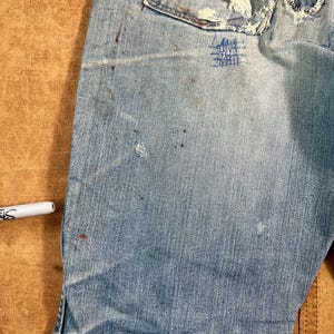 May include: Close-up of light blue denim jeans with visible wear and tear. The jeans have a distressed look with patches, frayed edges, and paint splatters. A white marker is visible in the image.