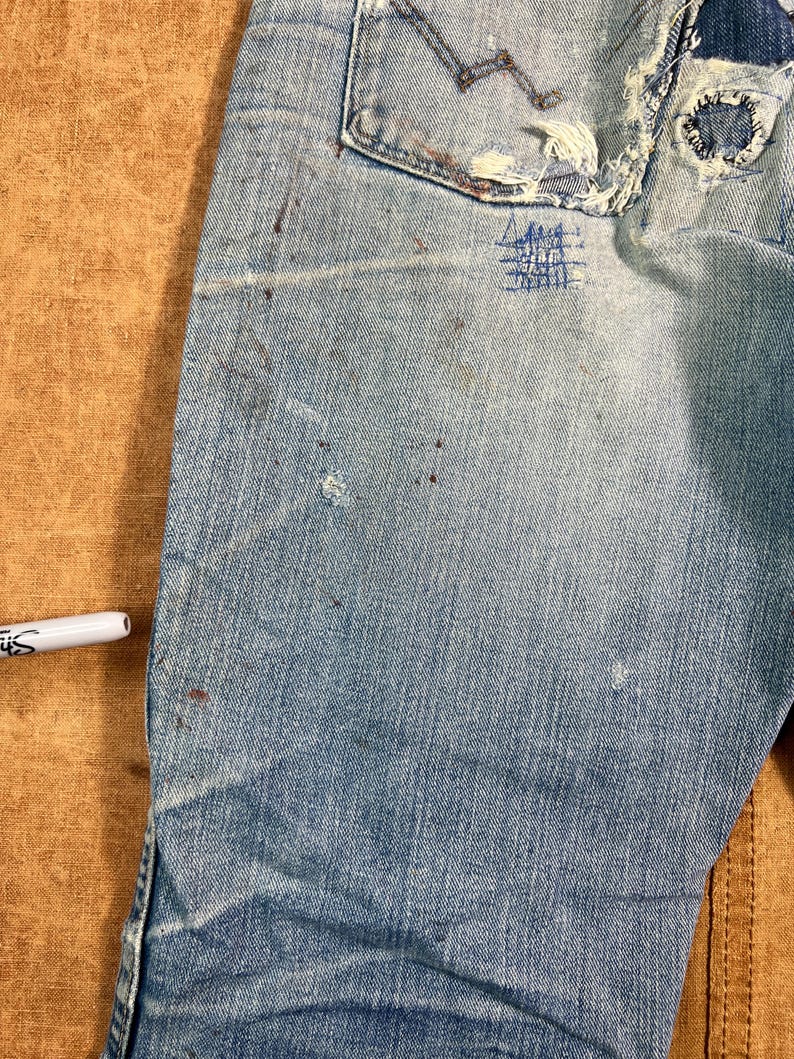 May include: Close-up of light blue denim jeans with visible wear and tear. The jeans have a distressed look with patches, frayed edges, and paint splatters. A white marker is visible in the image.