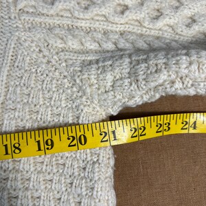 May include: Cream-colored knitted sweater with intricate cable and textured patterns. A yellow measuring tape shows a section of the sweater is approximately 6 inches wide. The sweater's design includes diamond and braided details.