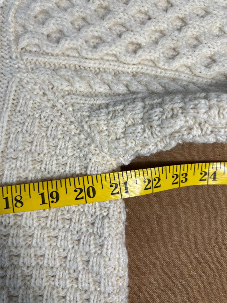 May include: Cream-colored knitted sweater with intricate cable and textured patterns. A yellow measuring tape shows a section of the sweater is approximately 6 inches wide. The sweater's design includes diamond and braided details.