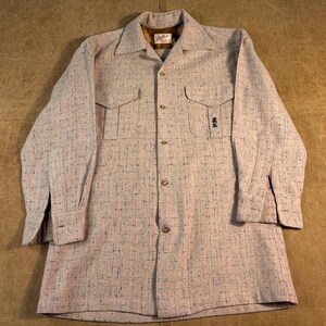 May include: A long-sleeved button-down shirt in a light beige fabric with a dark brown cross-hatch pattern. The shirt features a pointed collar, two chest pockets with flaps, and a small dark embroidered emblem on the left pocket. The inner collar label reads "Jack Lord".
