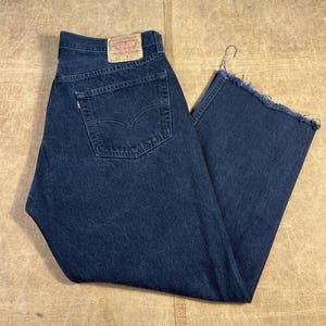 May include: Dark blue denim jeans with a classic five-pocket design. The jeans feature a red Levi's tag on the back pocket and a raw hem. The jeans are folded and displayed on a light brown surface.