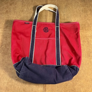 May include: A red canvas tote bag with navy blue accents and a white handle. The bag features a front pocket and a monogram in navy blue. The bottom of the bag is navy blue.