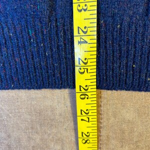 May include: Close-up of a blue knit fabric with flecks of red, green, and yellow. A yellow measuring tape is placed vertically along the fabric, showing measurements from 23 to 30 inches.