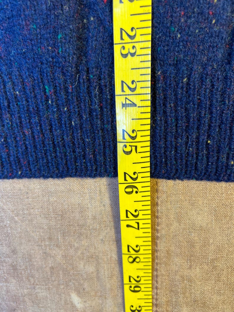May include: Close-up of a blue knit fabric with flecks of red, green, and yellow. A yellow measuring tape is placed vertically along the fabric, showing measurements from 23 to 30 inches.
