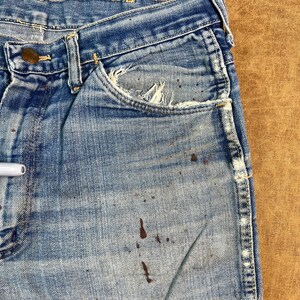 May include: Close-up of vintage blue denim jeans with a worn appearance. The jeans show signs of wear and tear, including distressed pockets and brown paint splatters. The jeans have a classic five-pocket design and a button closure.