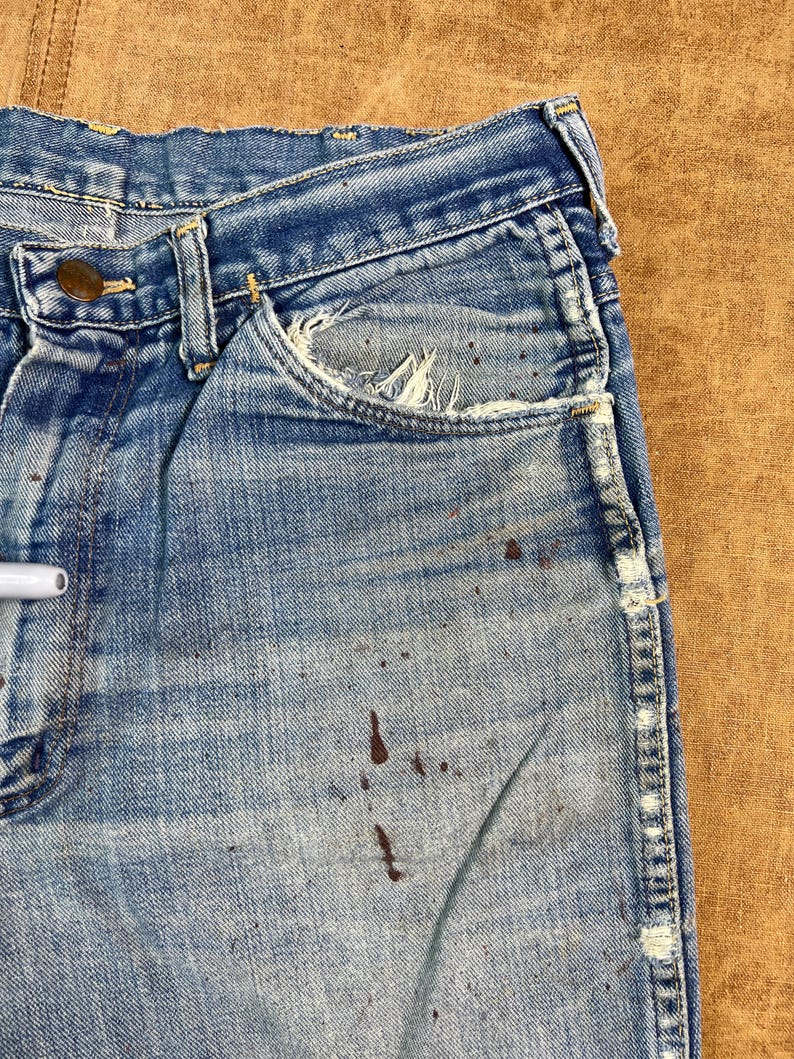 May include: Close-up of vintage blue denim jeans with a worn appearance. The jeans show signs of wear and tear, including distressed pockets and brown paint splatters. The jeans have a classic five-pocket design and a button closure.